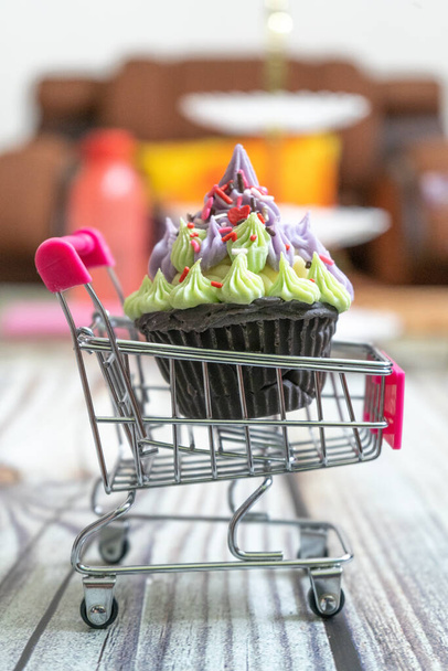 Beautifully iced cupcake with purple frosting and black chocolate base on a shopping cart on a wooden floor with blurred out background - Foto, Imagem