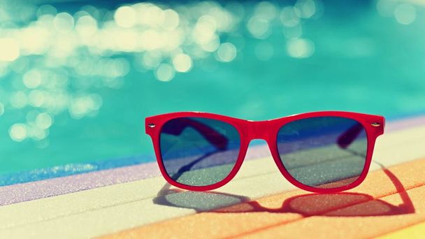 Zomer achtergrond. Concept voor zomer en vakantie. Rode zonnebril bij het zwembad op de achtergrond met blauw water. - Foto, afbeelding
