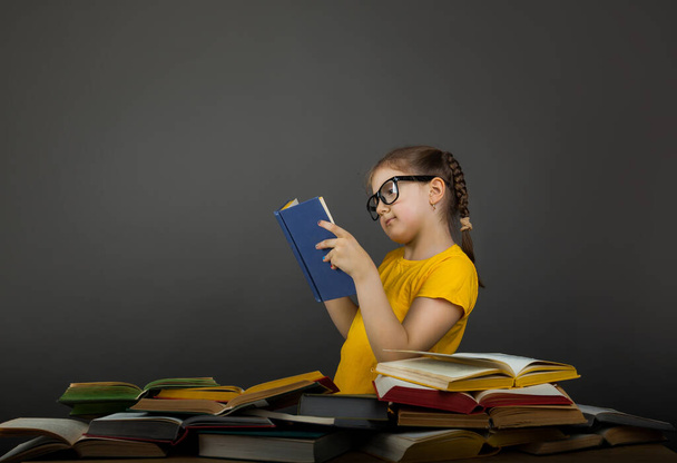 Nieuwsgierig klein meisje in geel t-shirt ziet er geweldig uit kijkend naar boeken, op grijze achtergrond - Foto, afbeelding