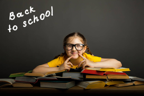 Back to school concept. Happy schoolgirl in glasses wearing a yellow t-shirt, at the black chalkboard in classroom. Chalk inscription on blackboard Back to school. - Fotó, kép