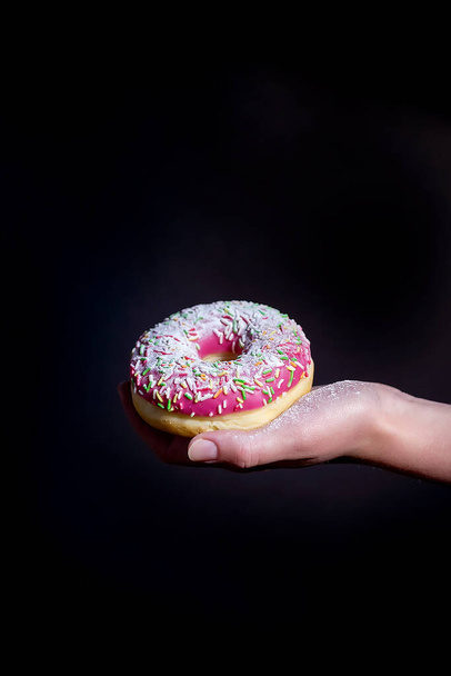 Donut on a dark background. Donut with pink frosting. The cook's hand holds a pink doughnut. Selective focus. Close up. - Foto, Imagem