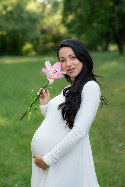 Donna incinta in posa in un abito bianco su uno sfondo di alberi verdi. - Foto, immagini