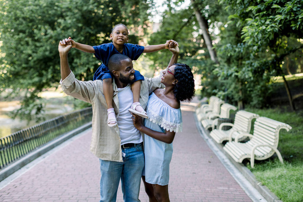 Family in nature in park. Handsome African American man father carrying his little daughter on shoulders, looking at his pretty smiling wife and enjoying walk outdoors - Foto, immagini