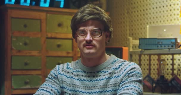 Portrait shot of Caucasian male goofy nerd in glasses with mustache sitting at desk in retro room and looking silly. Funny man with stupid face looking at camera. Vintage style of 80s or 70s. - Footage, Video