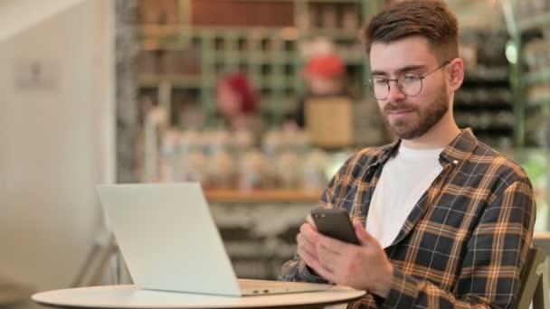 Hombre joven con el ordenador portátil hablando en el teléfono inteligente en Café
  - Metraje, vídeo