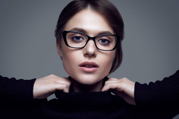 Porträt der netten Studentin mit schwarzem Rollkragenpullover und stylischer Brille posiert auf grauem Hintergrund im Studio - Foto, Bild