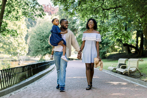 Full length portrait of adorable african family of three, father, mother and child, walking in the park. Handsome father holds his daughter in arms and holds hand of his charming wife - Foto, immagini