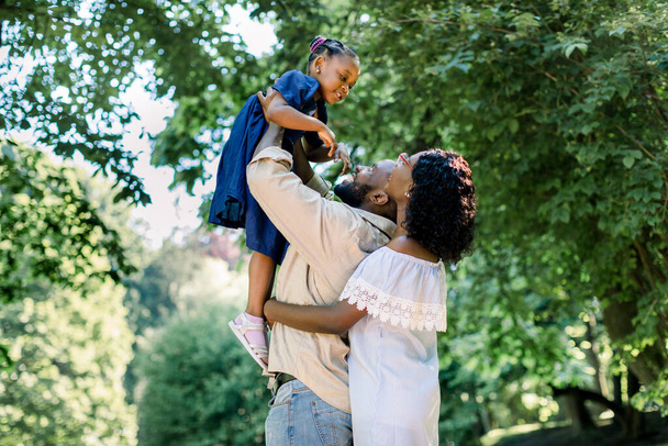 Summer shot of happy african family playing in the park. Family portrait with happy people smiling at the park, father lifts the cute girl child up. Lifestyle and happy family day concept - 写真・画像