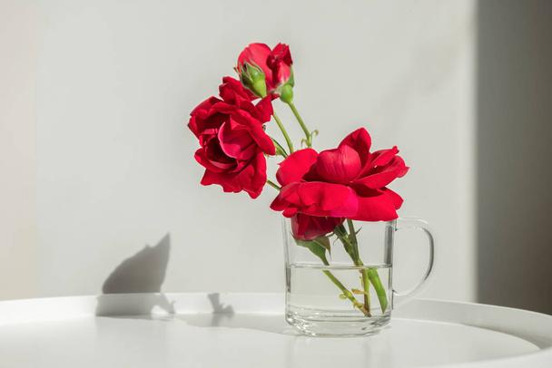 Интерьер, домашний декор. Красивые красные розы в стакане на белом столе
 - Фото, изображение