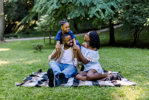 Happy African family of three having picnic in park on summer day. Young family outdoors in a park, sitting on a checkered blanket. Child girl is piggybacking on her daddy - Fotó, kép