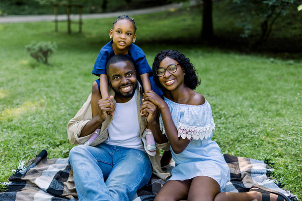 Adorable young African family with cute little daughter, having their weekend time in a city park. Image of cheerful family looking at the camera while picnicking in the park - Фото, изображение