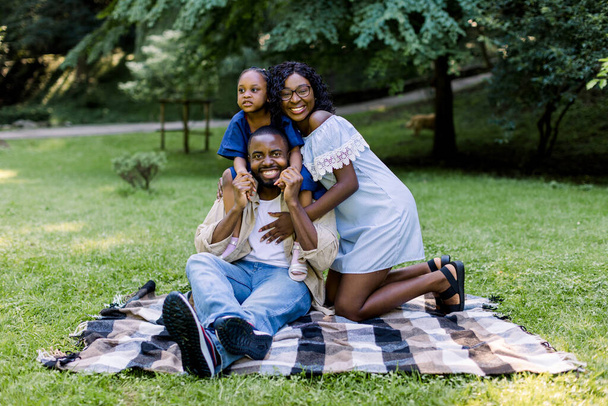 Family picnic in the park. Happy African family with child girl, wearing casual stylish clothes, sitting on checkered plaid in park and smiling to camera. Happy family concept. - Фото, зображення