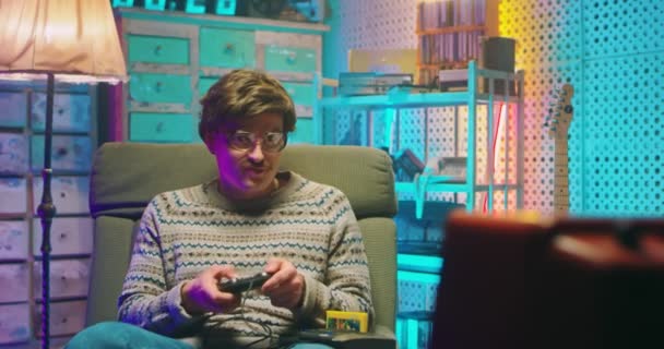 Grappige Kaukasische jongeman nerd in bril zittend voor vintage tv monitor en videogame spelend thuis met joystick. Mannelijke dwaze gek spelen videogames. Retro stijl van de jaren 80. Gamer uit de jaren 90. - Video