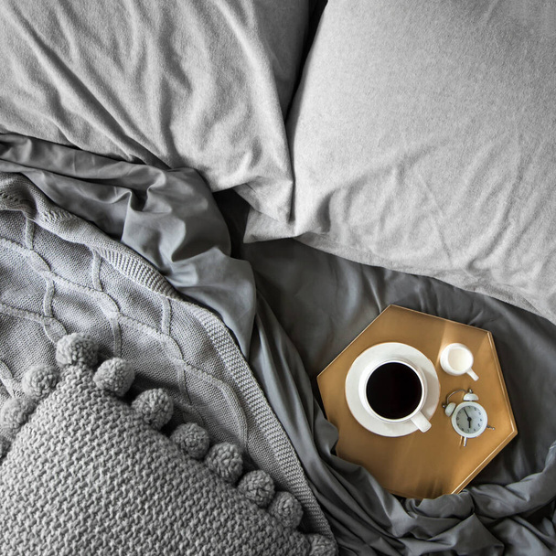 Morning breakfast in bed. Gray bedding. Golden tray in the nordic style. White cup with black coffee and a milkman with milk. Flowers in a gray vase. An alarm clock is set at seven in the morning. - Foto, imagen