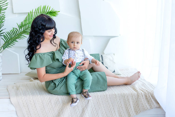 krásná mladá matka s dítětem v náručí sedí v jasném pokoji v zelených šatech, matka a syn, šťastný rodinný koncept, Den matek. - Fotografie, Obrázek