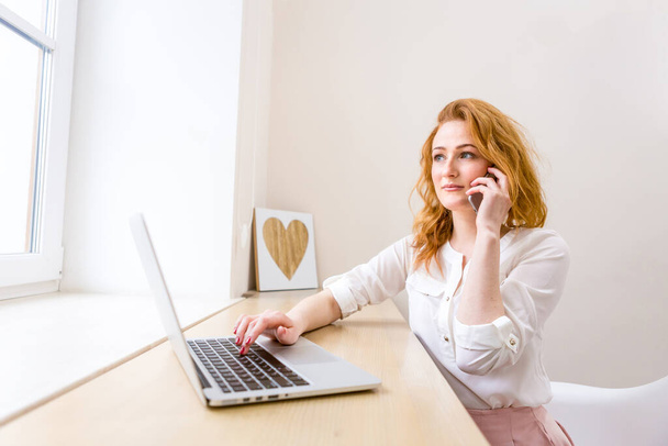 Bilgisayarlı ve akıllı telefonlu bir masada çalışan profesyonel bir iş kadını telefon görüşmesinde gülümsüyor, ofis içinde. Çalışan kadın yaşam tarzı. İş kadını Telefon görüşmesi yapıyor - Fotoğraf, Görsel
