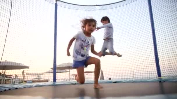 Šťastní sourozenci s úsměvem děti skákání na trampolíně na pláži při západu slunce. Sportovní děti se baví. Šťastné dětství a aktivní životní styl koncept. - Záběry, video