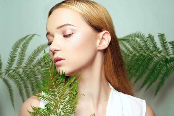 Girl in tropical leaves enjoys eyes closed. Portrait of a beautiful blonde with perfect clean skin. Pink makeup beauty photo, big lips, natural cosmetics - Zdjęcie, obraz