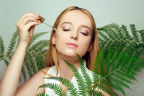 Girl on the background of tropical leaves natural cosmetics and medicines. The model holds a bottle and oil for the face and applies it to the skin. Bottle with a pipette. - Φωτογραφία, εικόνα
