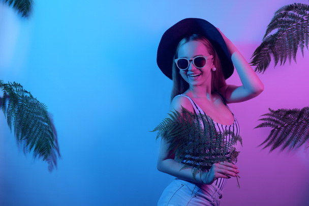 Egy lány trópusi levelek hátterében. Modell neon fekete kalapban fürdőruha napszemüveg és rövidnadrág. Tánc, klub, beach party, divat, nyári zene. - Fotó, kép
