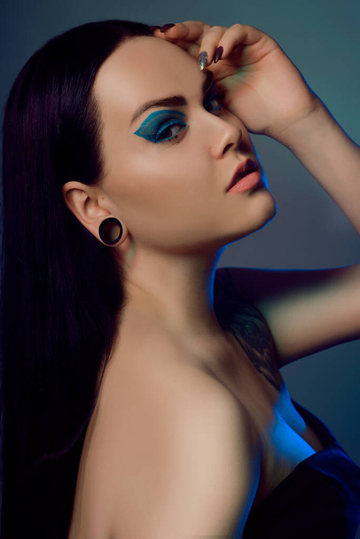 Brunette girl with tunnels in her ears in a beautiful blue light. Woman with blue make-up, fashionable blue arrows over the eyelid. Insightful brown eyes. Model with beautiful hair and professional makeup. - Foto, Bild
