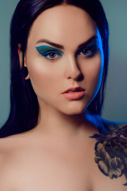 Μελαχρινή κοπέλα με ένα τατουάζ λουλούδι στον ώμο της και σήραγγες στα αυτιά της σε όμορφο μπλε φως. Γυναίκα με μπλε μακιγιάζ, μοντέρνα μπλε βέλη πάνω από το βλέφαρο. - Φωτογραφία, εικόνα