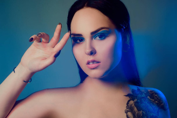 Brunette girl with a flower tattoo on her shoulder and tunnels in her ears in beautiful blue light. Woman with blue make-up, fashionable blue arrows over the eyelid. - Foto, afbeelding