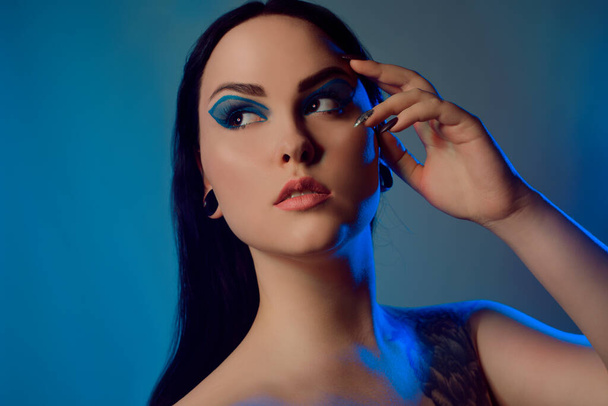 Brunette meisje met een bloem tatoeage op haar schouder en tunnels in haar oren in prachtig blauw licht. Vrouw met blauwe make-up, modieuze blauwe pijlen over het ooglid. - Foto, afbeelding