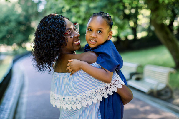 Beautiful family portrait at the summer sunny park of happy african mother and daughter together, smiling and enjoying walk. Pretty mom holds her kid girl on hands and looks at the baby. - Foto, Imagen