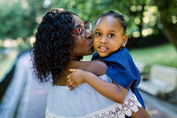 Close up portrait of little cute smiling African child girl in blue dress, looking at camera, while hugging pretty mother, kissing her in the cheek. African mother and daughter together in park - Foto, Bild