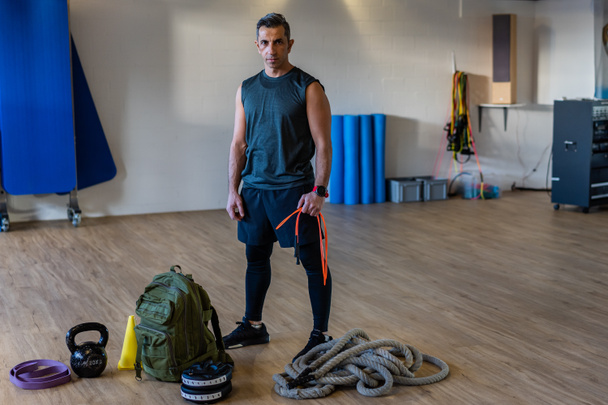 Motivated boot camp instructor stands with gym equipment in gym hall. Dumbbells, rope, sandbag on wooden floor. Portrait of lebanese instructor for gym concept. - Fotografie, Obrázek