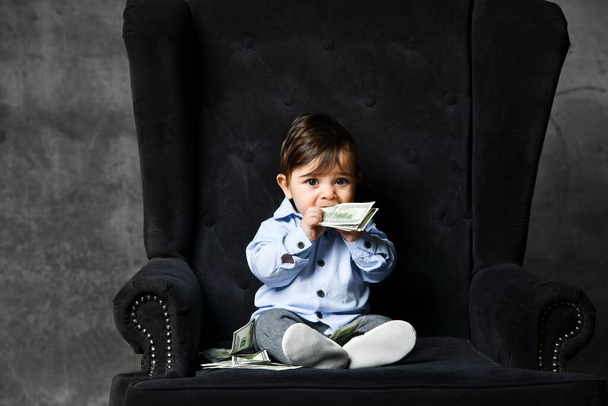 Toddler in blue shirt, gray pants, booties. He holding money, chewing it, sitting in black armchair on gray background. Close up - Foto, Imagen