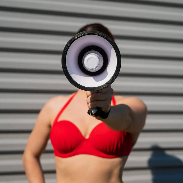 Close-up portrait of a woman in a red bikini holds a megaphone on the background of the face. A girl in a swimsuit makes an announcement on the speaker against the background of a steel sheet. - Φωτογραφία, εικόνα