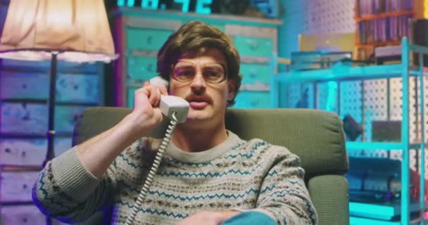 Kaukasische jongeman nerd in bril en met snor zittend in retro stijl huis en pratend op bedraad vintage telefoon van de jaren 80. Boos gestresste man aan de telefoon. Goofy jongen van de 90. - Video
