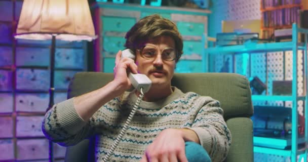 Kaukasische jongeman nerd in bril en met snor zittend in retro stijl huis en pratend op bedraad vintage telefoon van de jaren 80. Boos gestresste man spreekt over de telefoon tijdens het bellen. Goofy jongen van de 90 - Video