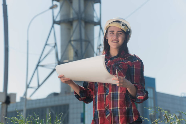 Portrait of a young beautiful girl in a protective white helmet, engineer woman holds a paper sketch in her hand on a background of a construction site. The concept is to copy space on the side - Photo, Image