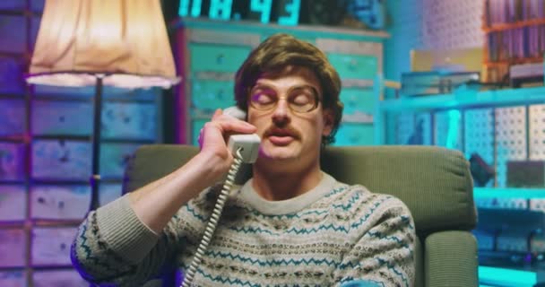 Kaukasische jongeman nerd in bril en met snor zittend in retro stijl huis en pratend op bedraad vintage telefoon van de jaren 80. Boze man met telefoontjes en zetten. Goofy jongen van de 90 - Video