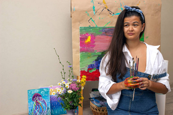 Chica sosteniendo sucios pinceles de pintura en las manos con la imagen en el fondo. El artista terminó la pintura abstracta en su taller
 - Foto, imagen