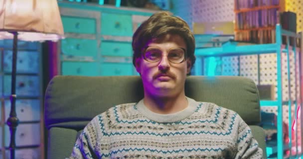 Portrait of young Caucasian man nerd in glasses and with mustache sitting in armchair in retro interior room and looking seriously to camera. Close up of male goofy from 80s. Vintage style. - Footage, Video