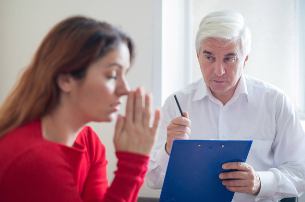 A frustrated crying woman holds palms in her face at a session with a male psychologist. Mature gray-haired psychotherapist talking to a female patient with depression and neurosis. Unbalanced psyche. - Foto, imagen