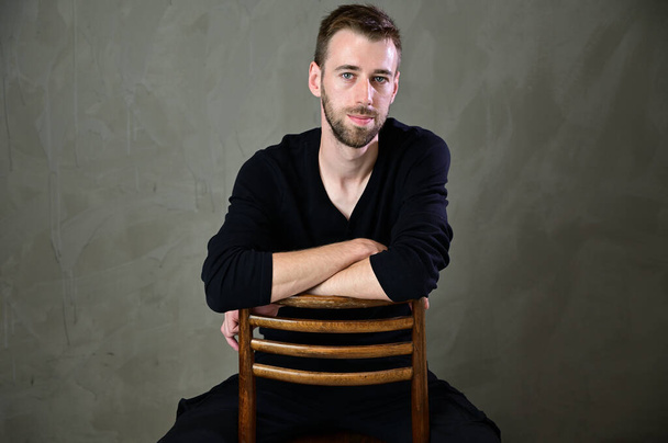 Stylish vertical studio portrait of a caucasian young man with a beard sitting on a chair on a gray background - Foto, imagen