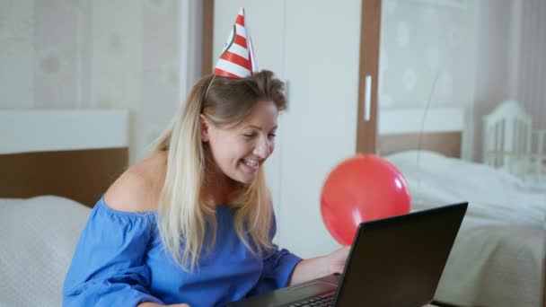 narozeniny on-line, radostná usměvavá žena s čepicí a míčky blahopřeje své kamarádce k dovolené s pomocí moderních technologií pomocí webové kamery na notebooku v důsledku sociální distancování během pandemie a - Záběry, video