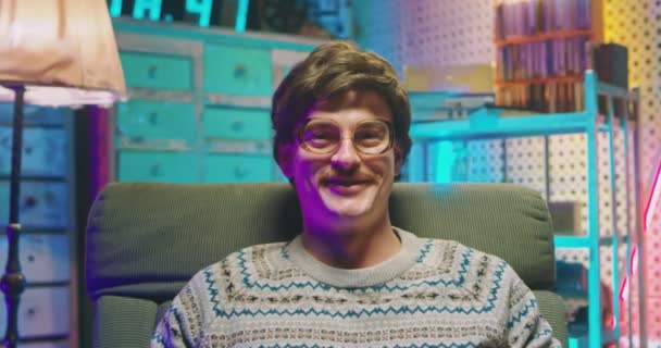 Portrait of young handsome Caucasian man nerd in glasses and with mustache sitting in armchair in retro interior room and smiling joyfully to camera. Close up of happy male goofy from 80s. Vintage. - Footage, Video