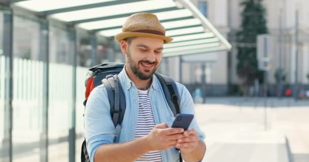 Fiatal jóképű kaukázusi férfi kalapban, hátizsákkal sms-ezik az okostelefonon, és vidáman mosolyog a vasútállomáson. Vonzó férfi utazó lehallgatás és görgetés mobiltelefonon a buszmegállóban. - Felvétel, videó