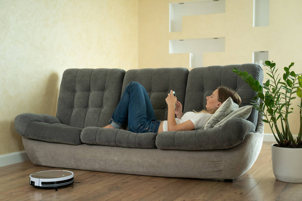 Attractive girl using smartphone while robotic vacuum cleaner cleaning house - Foto, Bild