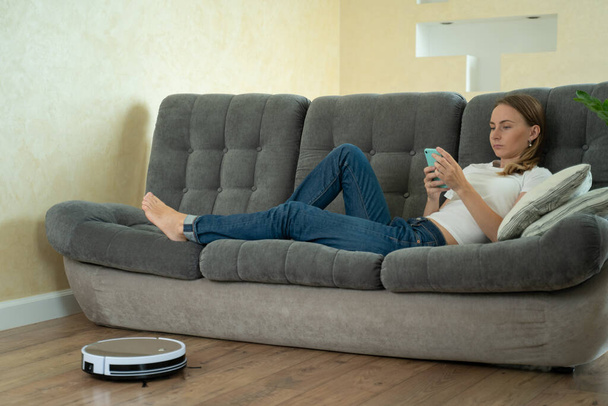Attractive girl using smartphone while robotic vacuum cleaner cleaning house - Fotoğraf, Görsel