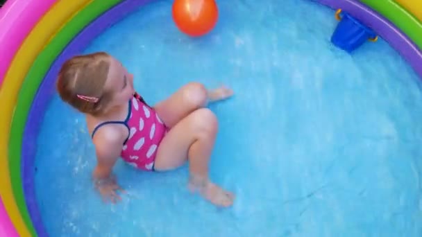 Jeune fille en maillot de bain rose jouant dans la piscine gonflable arc-en-ciel colorée sur une arrière-cour. Été domestique style de vie actif nager pour les enfants, plaisir à la maison loisirs nautiques pour les enfants, le temps chaud 4K vidéo - Séquence, vidéo