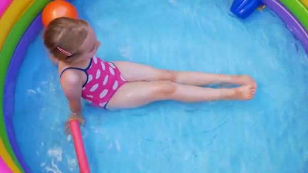 Giovane ragazza in costume da bagno rosa che gioca in colorata piscina gonfiabile arcobaleno su un cortile. Estate domestico stile di vita attivo nuotare per i bambini, divertimento a casa acqua per il tempo libero per i bambini, caldo tempo 4K video - Filmati, video