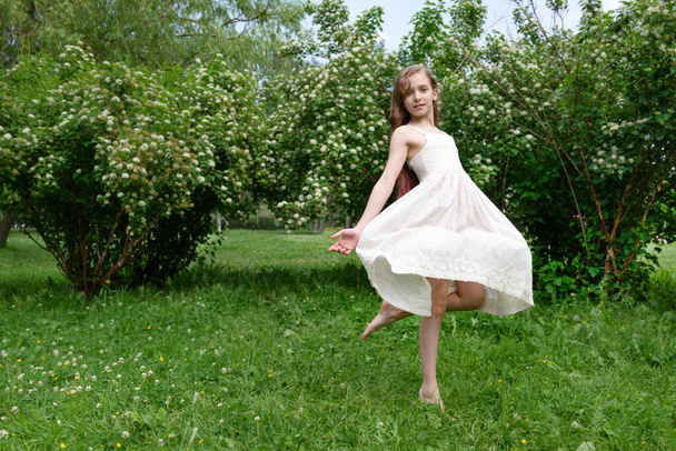 1 white beautiful girl 11 years old with long hair in a white dress spinning on the grass in a clearing against the background of greenery in the garden - Photo, image