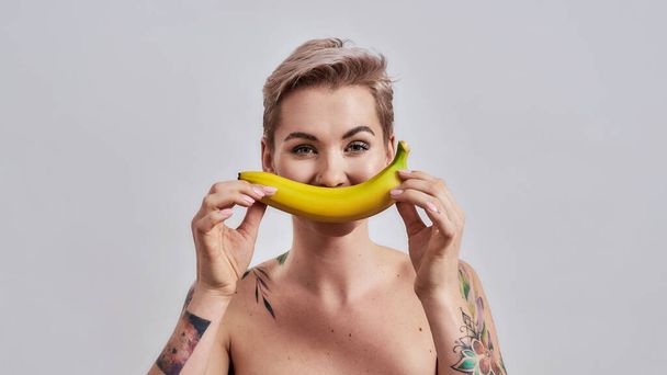 Cute and Fruit. Attractive tattooed woman with pierced nose and short hair holding banana as her smile isolated over grey background - Foto, afbeelding